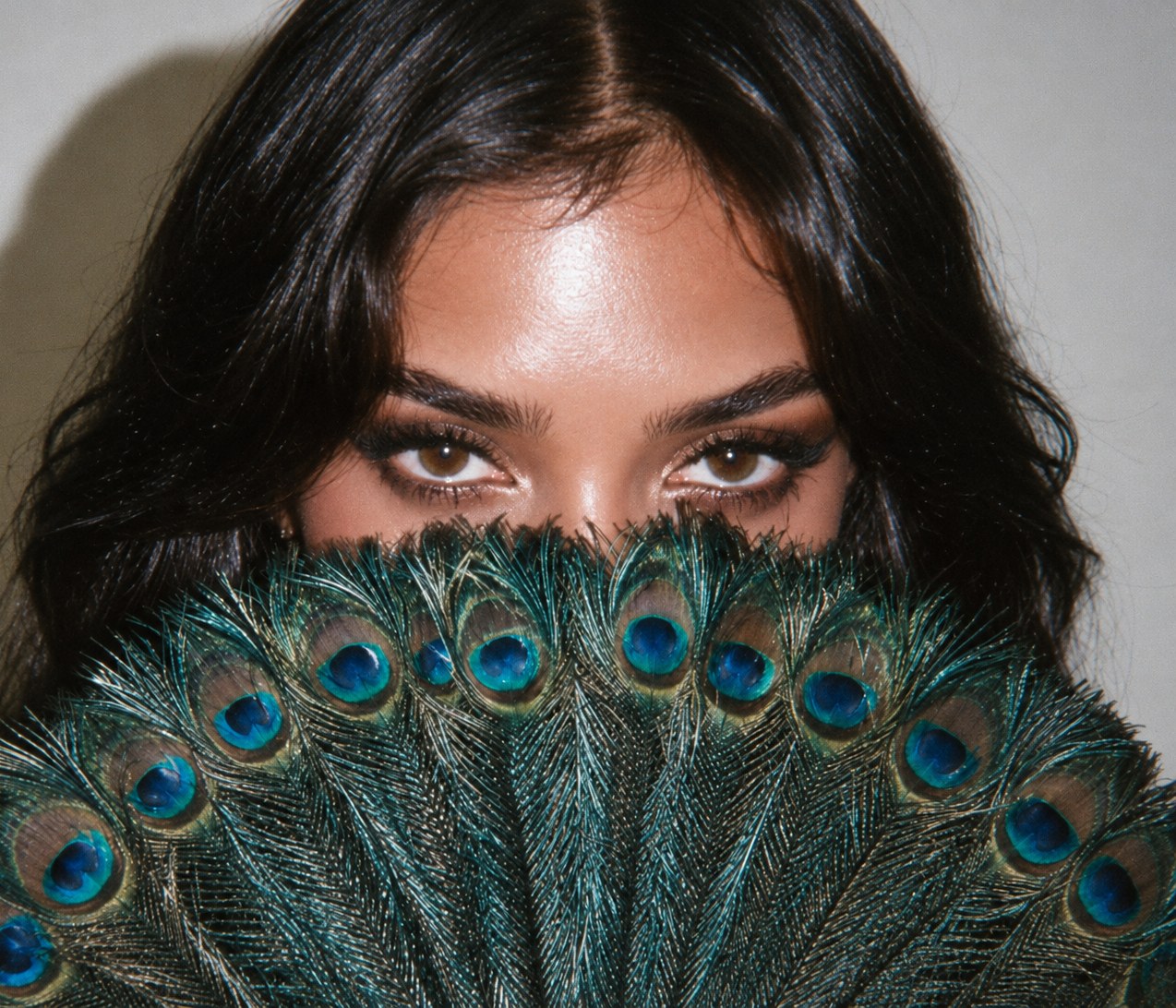 A woman in glossy evening makeup looks over a fan of peacock feathers held across her face